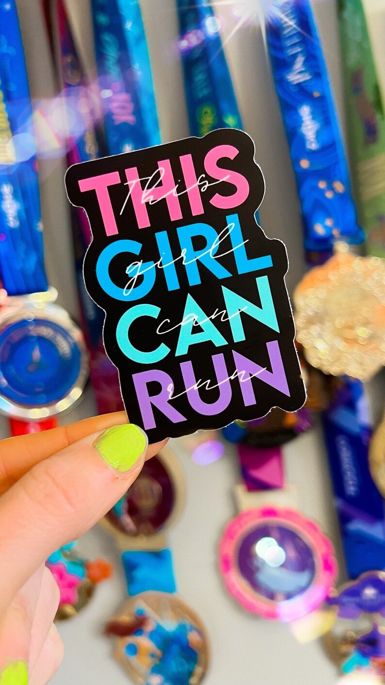 May include: A black sticker with the text "This girl can run" in pink, blue, and purple letters. The sticker is held in a hand with green nail polish.