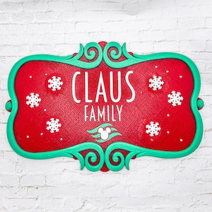 May include: A rectangular red sign with a decorative teal border featuring the white text CLAUS FAMILY. The sign is decorated with white snowflake cutouts and a small white icon of a mouse head centered above three teal wavy lines.