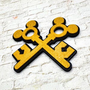 May include: Two gold-colored keys crossed over each other, with a black outline. The keys are set against a white brick wall. The keys have a textured surface and a decorative design.