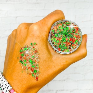 May include: A small jar of green and red glitter with a silver base. The glitter is scattered on a hand.