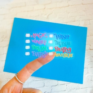 May include: A blue card with a clear sticker featuring colorful text that reads "Magic, Wish, Wonder, Treasure, Dream, Destiny, Fantasy, Adventure." The sticker has check boxes next to each word. A finger with a silver ring is in the foreground.