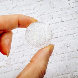 May include: A small clear container filled with iridescent glitter. The glitter is a mix of white, silver, and rainbow colors.