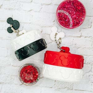 May include: Two cylindrical containers with white and black or red bands featuring embossed mouse head silhouettes. Each includes a small mouse-shaped charm on a metal chain. Two small circular jars filled with pink and red glitter are also present.