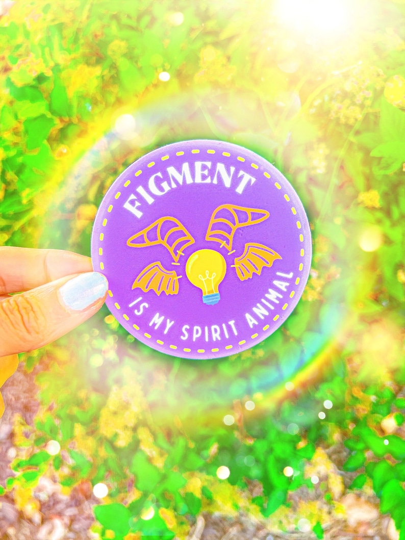 Puede incluir: Una pegatina redonda morada con el texto "FIGMENT IS MY SPIRIT ANIMAL" en blanco. La pegatina presenta una bombilla amarilla con alas y dise&ntilde;os de alas amarillas. La pegatina est&aacute; sobre un fondo verde y amarillo borroso.