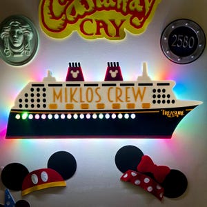 May include: A decorative cruise ship sign with the text "MIKLOS CREW" and "Treasure". The ship has red and black accents, with Mickey Mouse silhouettes on the smokestacks. The sign is illuminated with colorful LED lights.