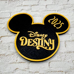 May include: Black and gold Mickey Mouse-shaped sign with the words "Disney Destiny" and the year "2025". The sign has a textured surface and is set against a white brick wall.
