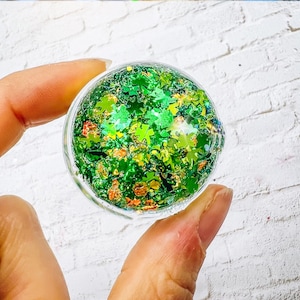 May include: A clear, round container filled with green and gold glitter in the shape of shamrocks. The glitter is suspended in a clear liquid. The container is held between two fingers against a white brick background.