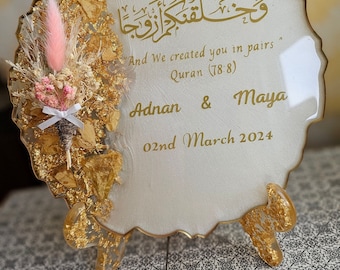 May include: A white circular resin plaque with gold leaf accents and a floral arrangement. The plaque features an Arabic inscription, "And We created you in pairs" Quran (78:8), and the names Adnan & Maya, along with the date 02nd March 2024.