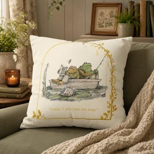 May include: A square off-white throw pillow featuring a printed illustration of two frogs in a small boat on water. One frog reads a book while the other fishes. A golden arch frame surrounds the scene with the text, Today, I will take life easy.
