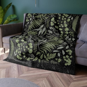 May include: A black throw blanket with a green botanical print featuring various leaves and ferns. The blanket is draped over a gray couch.