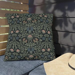 May include: Square decorative pillow with a dark background and a floral pattern in shades of green, pink, and blue. The pillow is placed on a blue cushion, with a straw hat in the foreground.
