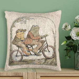 May include: Beige decorative pillow featuring a whimsical illustration of two frogs riding a bicycle. The frogs are dressed in vintage clothing, set against a backdrop of foliage within an arched frame. The pillow has a textured appearance.