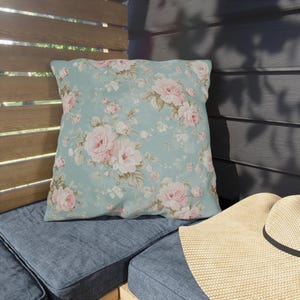 May include: Square throw pillow with a floral pattern of pink roses and green leaves on a light blue background. The pillow is placed on a blue cushion, with a straw hat visible in the foreground.
