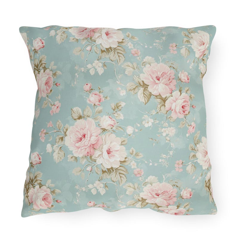 May include: Square throw pillow with a floral pattern. The pillow features a light blue background with a repeating pattern of pink and white roses and other flowers. The pillow has a soft, vintage aesthetic.
