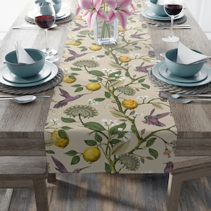 May include: A beige table runner with a floral pattern featuring lemons, white flowers, and purple hummingbirds. The runner is on a wooden table with place settings.