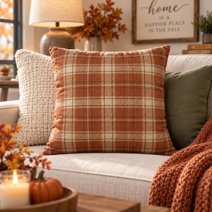 May include: A square throw pillow featuring a plaid pattern in shades of burnt orange, cream, and dark brown. The fabric has a visible woven texture, and the pillow is positioned on a light-colored sofa alongside other solid and textured pillows.