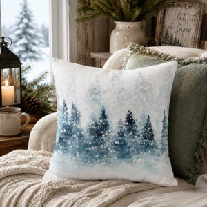 May include: A square throw pillow featuring a watercolor-style print of a snowy forest with deep blue and white pine trees. The design depicts a winter landscape with soft, blurred edges and white speckles representing falling snow across the fabric.
