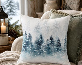 May include: A square throw pillow featuring a watercolor-style print of a snowy forest with deep blue and white pine trees. The design depicts a winter landscape with soft, blurred edges and white speckles representing falling snow across the fabric.