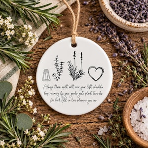 May include: A round white ceramic ornament hanging from a jute string. The front features black line art of a salt shaker, a rosemary sprig, a lavender stalk, and a heart. Text reads: Always throw spilt salt over your left shoulder keep rosemary by your garden gate plant lavender for luck fall in love wherever you can.