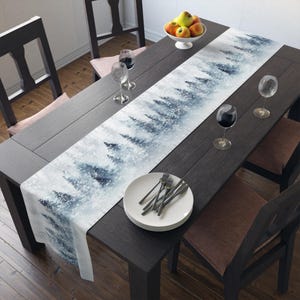 May include: A white table runner with a watercolor design of a snowy forest. The runner is placed on a dark wood table with white plates and silverware.