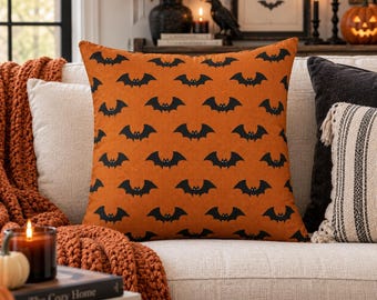 May include: A square throw pillow featuring a repeating pattern of black bats on a vibrant orange background. The fabric has a textured weave, and the design displays multiple rows of stylized bats with small white eyes across the entire surface.