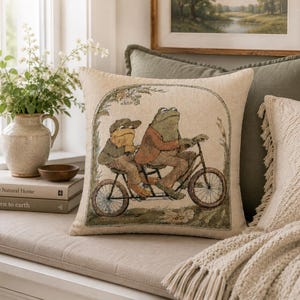 May include: A square throw pillow featuring a woven illustration of two anthropomorphic frogs riding a tandem bicycle. The scene is framed by an archway of foliage. The pillow has a light beige background with muted green and brown tones.