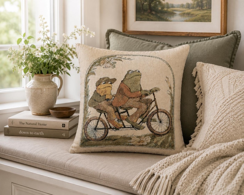 May include: A square throw pillow featuring a woven illustration of two anthropomorphic frogs riding a tandem bicycle. The scene is framed by an archway of foliage. The pillow has a light beige background with muted green and brown tones.