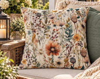 May include: A square throw pillow featuring a cream-colored background with a detailed botanical print of various wildflowers, dried grasses, and foliage in shades of orange, yellow, muted green, brown, and soft blue.