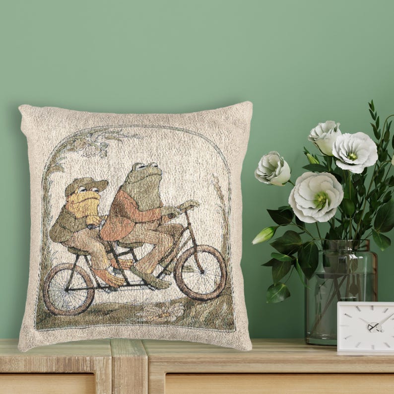 May include: A square throw pillow featuring a woven illustration of two frogs riding a tandem bicycle. The frogs are wearing jackets and hats, depicted in muted earth tones of brown, tan, and green against a light beige background with an arch.