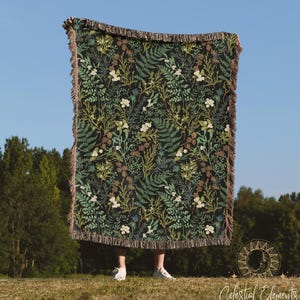 May include: A woven throw blanket with a dark green background and a floral pattern featuring ferns, leaves, and white flowers. The blanket has fringe on all sides.
