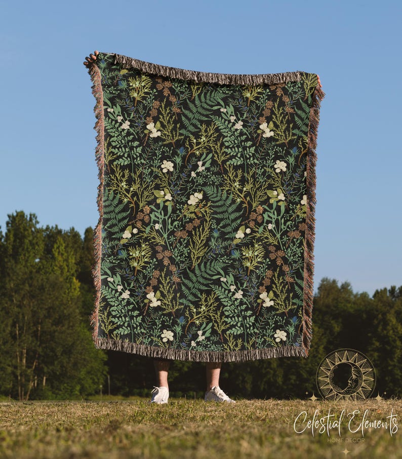 May include: A woven throw blanket with a dark green background and a floral pattern featuring ferns, leaves, and white flowers. The blanket has fringe on all sides.