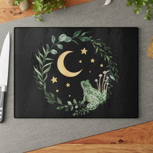 May include: A black cutting board with a green frog sitting on a wreath of green leaves. The frog is looking up at a yellow crescent moon with yellow stars in the background.