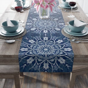 May include: A blue and white table runner with a snowflake design. The runner is on a wooden table with two place settings on each side.