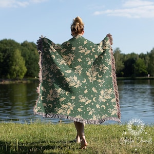 May include: A green woven throw blanket with a beige celestial pattern of moons, stars, and foliage. The blanket has a fringed edge and is held up by a person standing near a lake. The blanket is labeled "Celestial Elements".
