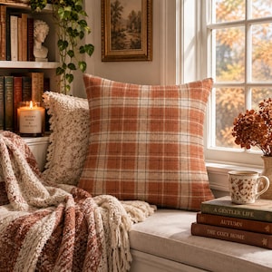 May include: A square throw pillow featuring a plaid pattern in shades of burnt orange, cream, and thin black lines. The fabric has a visible woven texture. The pillow is positioned near a window, accompanied by a cream-colored textured throw.