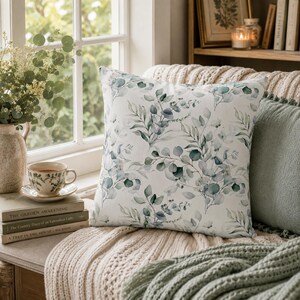May include: A square throw pillow featuring a white background with a repeating watercolor pattern of green and blue-toned eucalyptus leaves and branches. The botanical print covers the entire front surface of the pillow in a soft, organic arrangement.