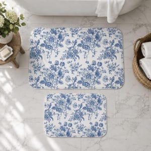 May include: A set of two rectangular bath mats featuring a white background with a repeating blue floral pattern. The design consists of intricate botanical illustrations of various flowers and leaves, creating a consistent visual theme across both mats.