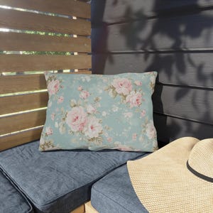 May include: A rectangular decorative pillow with a floral pattern of pink roses on a light blue background. The pillow is placed on a blue cushion, with a straw hat visible in the foreground. The scene is set outdoors.