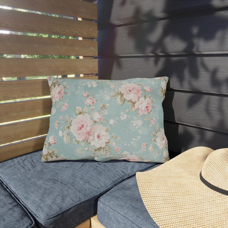 May include: A rectangular decorative pillow with a floral pattern of pink roses on a light blue background. The pillow is placed on a blue cushion, with a straw hat visible in the foreground. The scene is set outdoors.