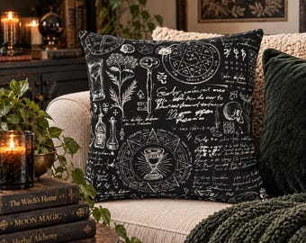 May include: A square throw pillow featuring a black fabric cover with white line art illustrations of an hourglass, a key, a chalice, a skull, botanical elements, geometric symbols, and handwritten script text covering the entire surface.