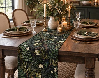 May include: A dark-colored table runner featuring a botanical print of green ferns, pine needles, and white berries. The runner is centered on a wooden dining table set with plates, napkins, glassware, and candles in a dining room setting.
