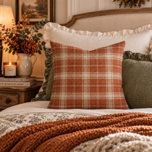 May include: A square throw pillow featuring a plaid pattern in shades of burnt orange, cream, and dark brown. The fabric has a visible woven texture, and the pillow is positioned centrally among other bedding items on a bed.