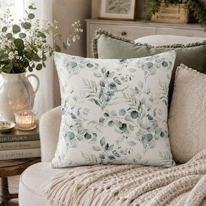 May include: A square throw pillow featuring a white background with a repeating watercolor pattern of green and blue-toned eucalyptus leaves and branches. The design displays various foliage shapes and stems arranged in a botanical print.