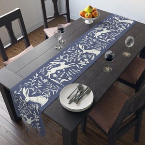 May include: A blue and white table runner with a repeating pattern of hares and floral designs. The runner is laid on a dark wood table with white plates and silverware.