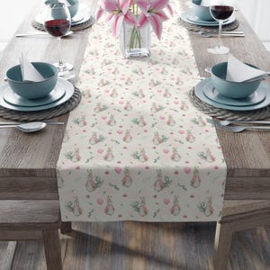 May include: A white table runner with a repeating pattern of pink bunnies, pink balloons, and green leaves. The runner is on a wooden table with two place settings on each side.