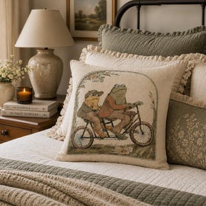 May include: A square throw pillow featuring a woven illustration of two anthropomorphic frogs riding a tandem bicycle. The scene is framed by a decorative archway on a light beige fabric background. The pillow has a textured, cream-colored fringe.