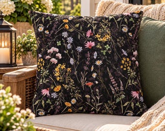 May include: A square throw pillow featuring a black background with a botanical print of various wildflowers in shades of yellow, pink, white, and light purple. The floral pattern includes daisies, lavender, and other small meadow plants.
