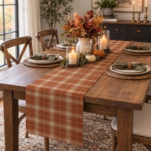 May include: A rectangular table runner featuring a burnt orange and cream plaid pattern. The runner is laid across a wooden dining table set with woven placemats, white plates, olive green cloth napkins, glass candle holders, and a floral centerpiece.