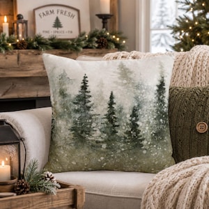 May include: A square throw pillow featuring a watercolor-style print of a dense forest with evergreen trees in shades of dark green and muted sage. The design includes a soft, speckled white overlay that mimics a light snowfall effect.