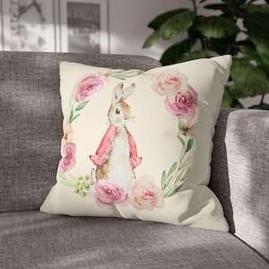 May include: A cream-colored throw pillow with a watercolor illustration of a rabbit wearing a pink jacket surrounded by pink flowers and green leaves.